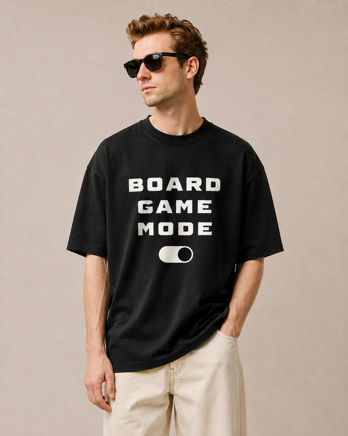 A man wearing a black graphic t-shirt with the text 'BOARD GAME MODE' and a toggle switch graphic in white, paired with beige shorts and black sunglasses, against a neutral beige background.